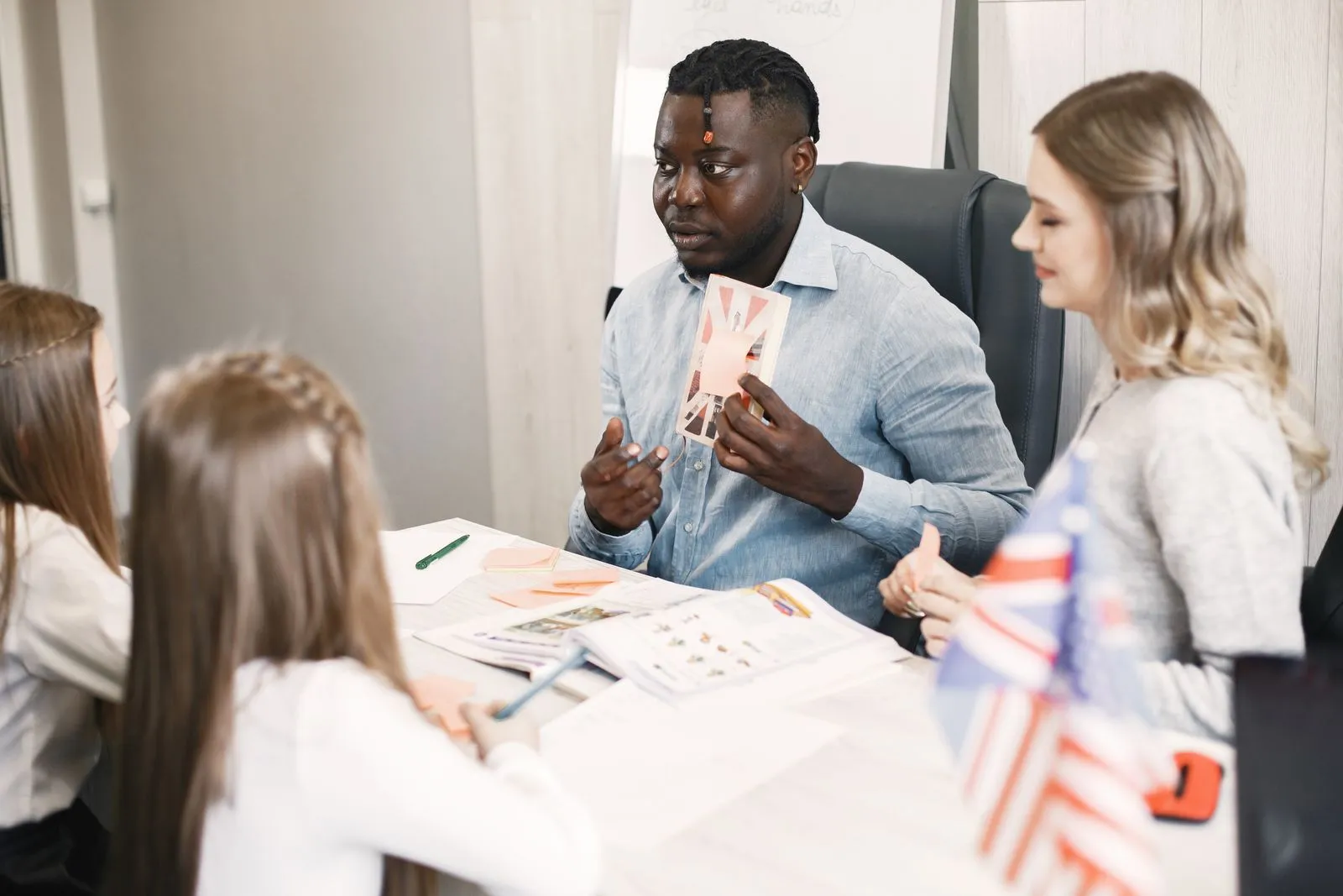 Professor de inglês conversando com alunas em volta de uma mesa com material de estudo