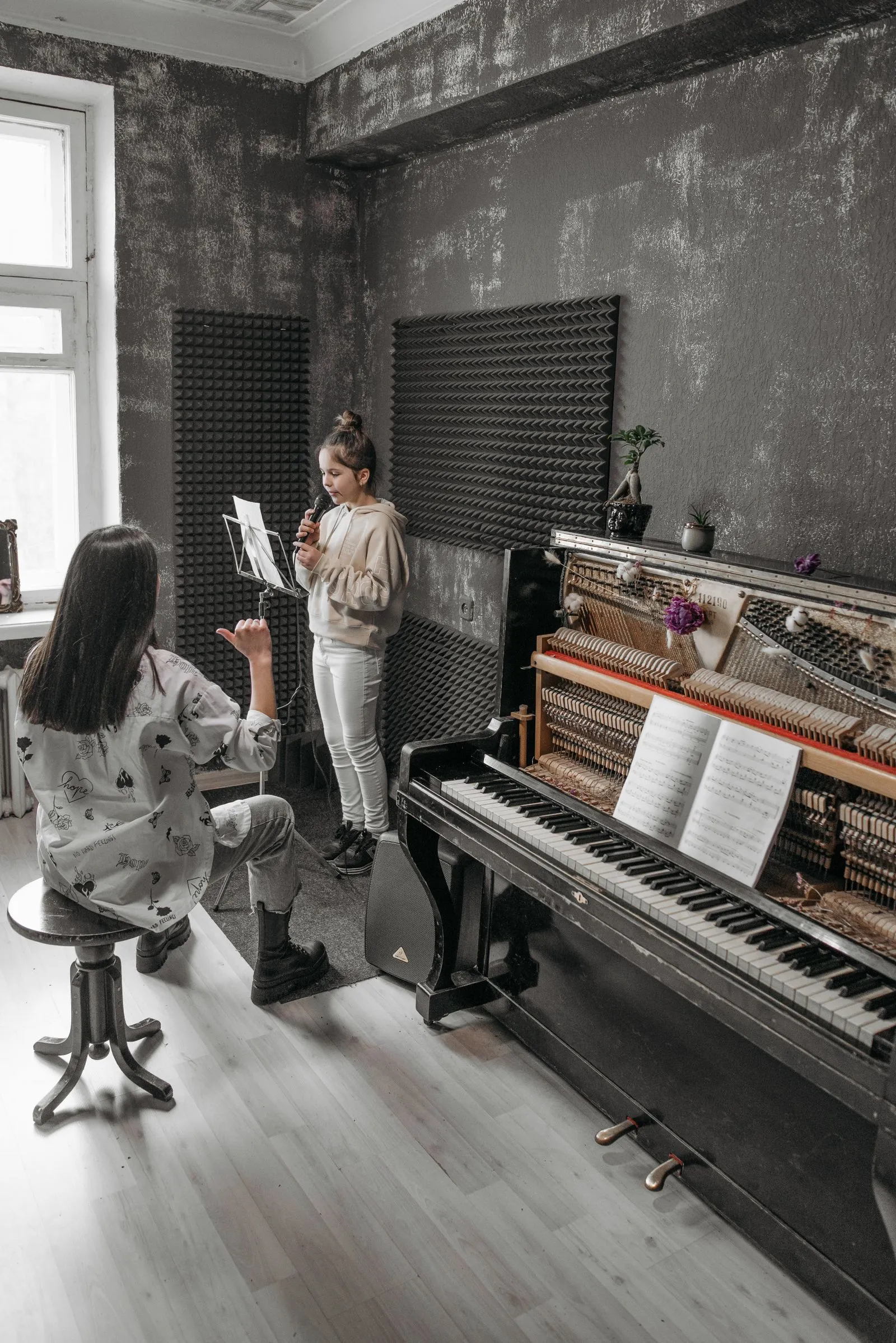 Menina canta ao microfone diante de uma estante de partitura, orientada pela professora, ao lado de um piano
