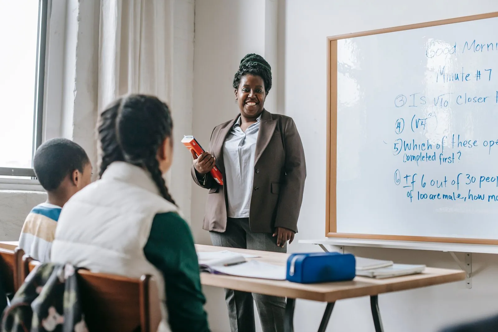 Professora sorridente em frente ao quadro branco conversando com alunos em sala de aula